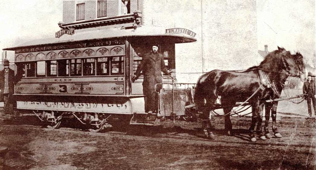 A horse-drawn streetcar on rails, with a conductor standing on the platform and two horses harnessed in front